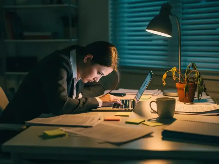 Exhausted businesswoman slumped over laptop at cluttered home office desk with coffee mug and wilted plant in evening light
