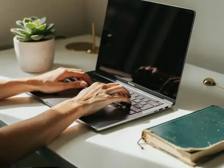 Woman's hands resting on open laptop at minimalist desk with succulent, notebook, and soft natural window light.