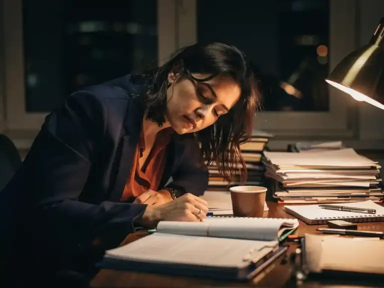 Exhausted professional woman slumped over a cluttered desk at night, surrounded by notebooks and scattered papers under warm lamplight.
