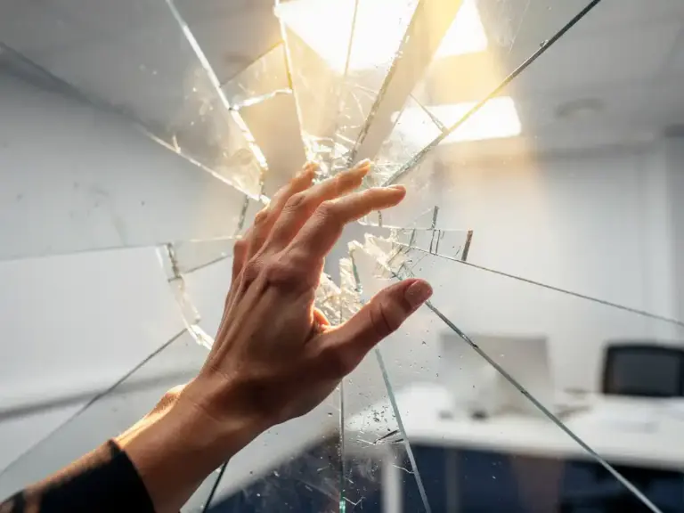 Woman's hand breaking through shattered frosted glass ceiling, golden light streaming through the burst, symbolizing career breakthrough in a white office setting.