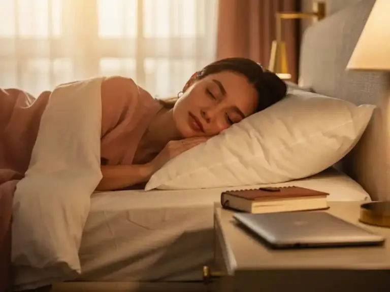 Professional woman sleeping peacefully on a white pillow, soft morning light illuminating a planner and laptop on the nightstand beside her.