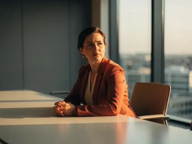 Professional woman seated alone at a minimalist conference table, hands folded calmly, warm afternoon light casting soft shadows across her thoughtful expression.