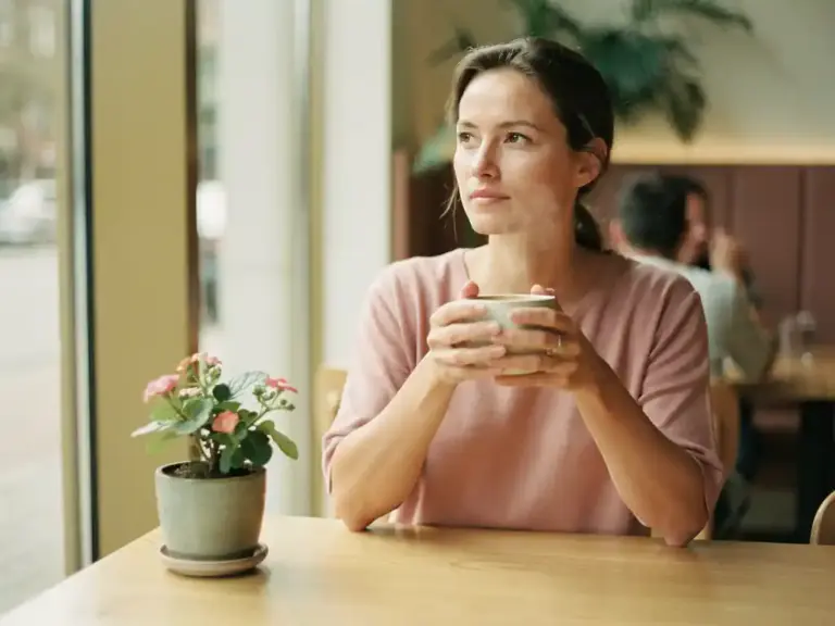 Woman sitting calmly at a sunlit café table, hands wrapped around a coffee cup, with a small blooming plant and soft morning light nearby.